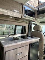 Inside an RV rental: a modern galley with a sink, microwave, induction cooktop, fridge, and wood-grain cabinets.