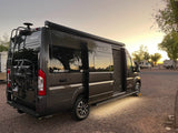 Dark grey Winnebago Travato RV rental shining at sunset in a gravel RV park. Features a ladder, bike rack, and illuminated side entry.