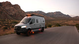 An RV rental, a grey Mercedes-Benz camper van, parked on a desert road with illuminated lights against rugged mountains at dusk.