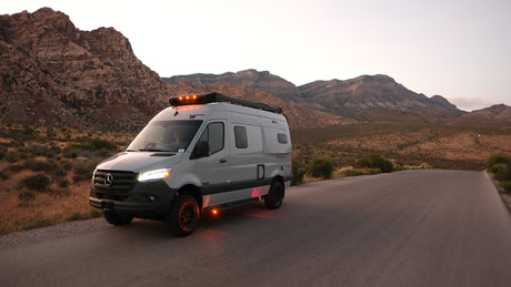 An RV rental, a grey Mercedes-Benz camper van, parked on a desert road with illuminated lights against rugged mountains at dusk.
