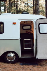 Cozy RV rental in the forest with an open door showing the interior kitchenette. Perfect for glamping.