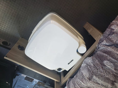 Portable Thetford toilet on a wooden platform in an RV rental, next to a fluffy blanket. Essential for van life.