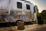 An Airstream RV rental on a wooden deck with two vibrant chairs, a tree stump, and a YETI cooler. Perfect for outdoor glamping.