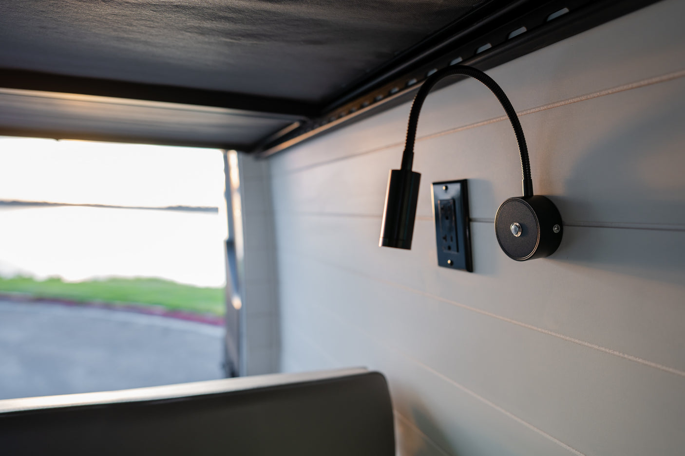 Inside an RV rental: black gooseneck lamps and an outlet on a paneled wall, with a serene lake view outside.