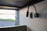 Inside an RV rental: black gooseneck lamps and an outlet on a paneled wall, with a serene lake view outside.