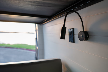 Inside an RV rental: black gooseneck lamps and an outlet on a paneled wall, with a serene lake view outside.