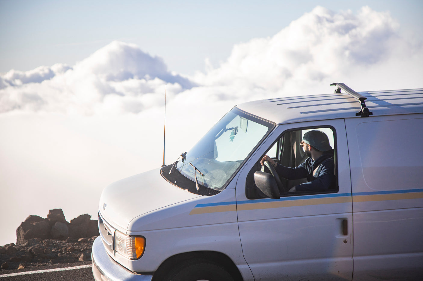 White RV rental van high on a mountain road, with a driver looking out over a sea of clouds.