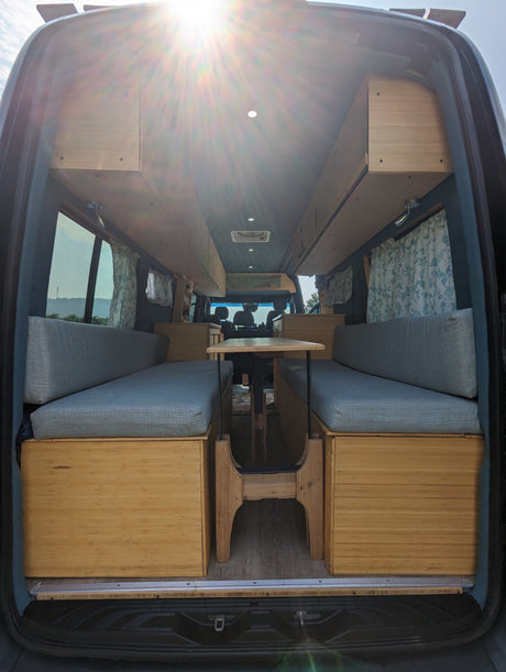Step inside this custom-built camper van interior, viewed from the rear. Features a dining area with wood benches, ideal for RV rental.