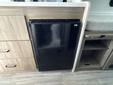 This RV rental interior features a compact black mini-fridge, light wood cabinetry with drawers, and adjacent open storage shelves.