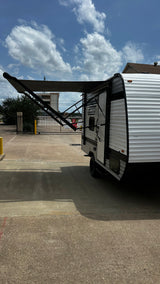 A white rv rental travel trailer with its dark awning extended on a sunny paved lot under a blue, cloudy sky.