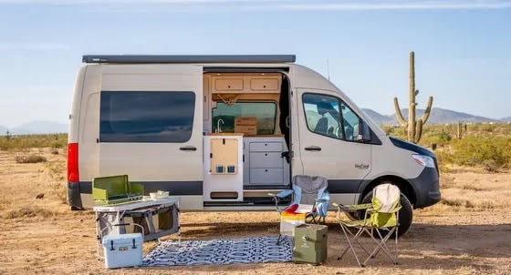 Explore the desert! This RV rental offers a fully equipped camper van with outdoor cooking and seating setup ready for adventure.|Tan camper van RV rental in a desert landscape with saguaro cacti and mountains under a clear blue sky.|Cozy RV rental interior with desert view. Features a functional kitchenette — photo 1