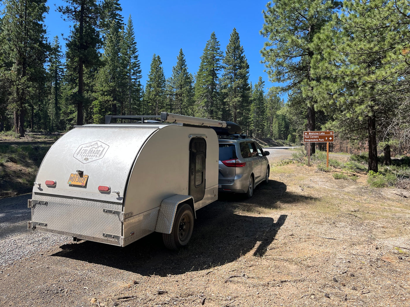 Perfect rv rental for forest adventures! Silver teardrop RV and minivan parked on a sunny dirt road in a pine forest.|RV rental: A silver minivan and light camper at a sunny forest campsite with a child's bike and chair. Ready for outdoor adventure!|An RV rental teardrop camper and minivan set up at a scenic mountain campsite.|Silver minivan with an RV rental teardrop trailer on a sunny road — photo 1