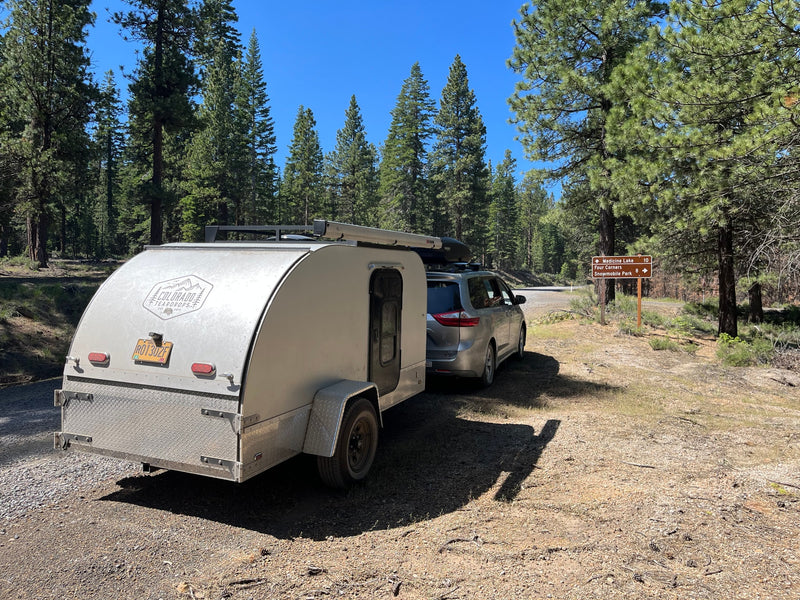 Perfect rv rental for forest adventures! Silver teardrop RV and minivan parked on a sunny dirt road in a pine forest.|RV rental: A silver minivan and light camper at a sunny forest campsite with a child's bike and chair. Ready for outdoor adventure!|An RV rental teardrop camper and minivan set up at a scenic mountain campsite.|Silver minivan with an RV rental teardrop trailer on a sunny road — photo 1