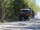 An RV rental, a brown VW van, kicks up dust on a dirt road, navigating a scenic pine forest.