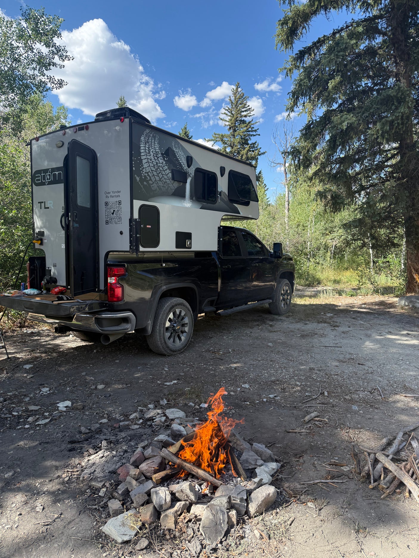 A modern truck camper, an RV rental, is set up on a black pickup truck at a rustic campsite with a bright campfire.