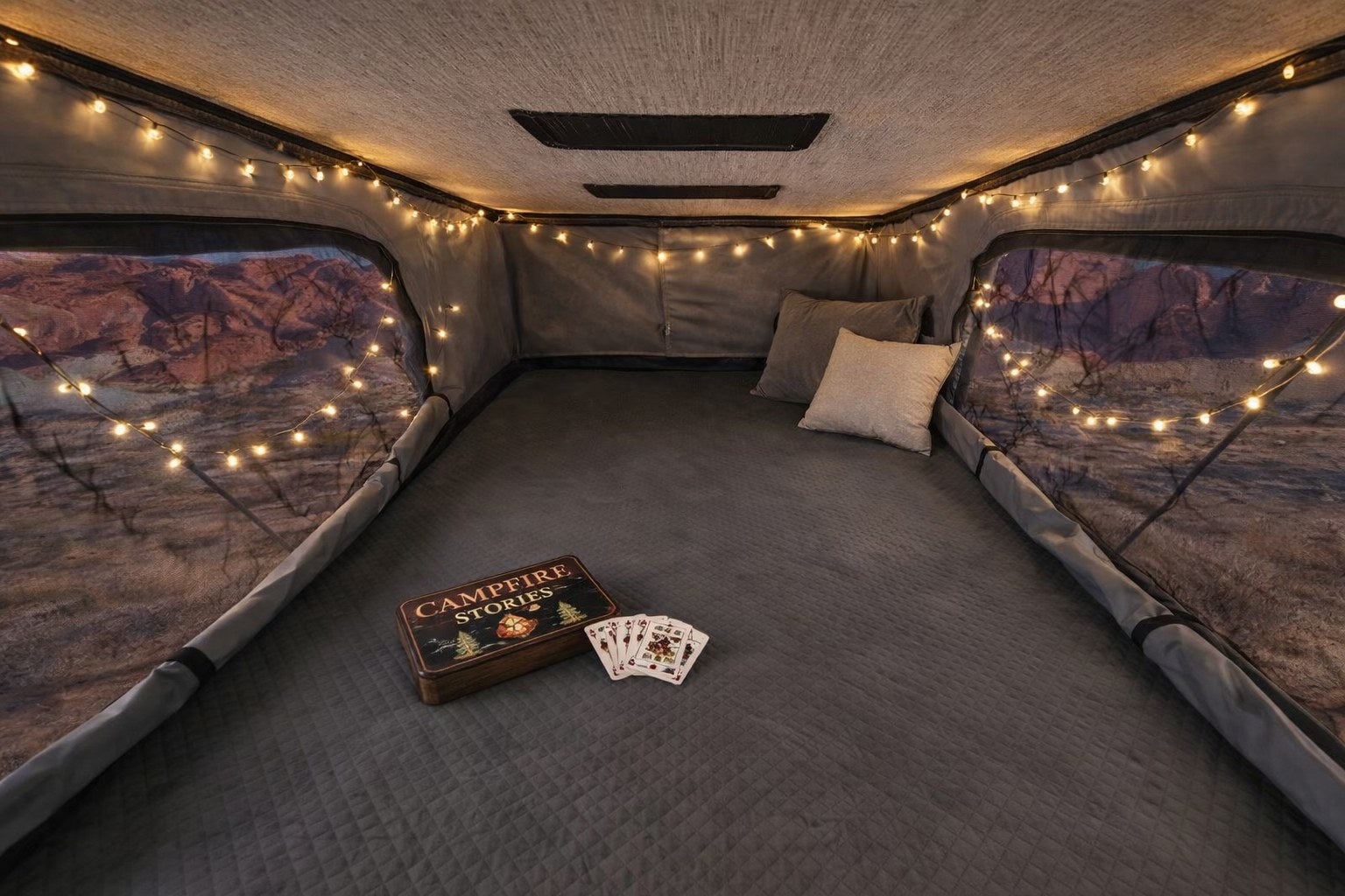 Cozy RV rental interior for glamping. String lights illuminate a comfy bed, scenic views, and Campfire Stories with playing cards.