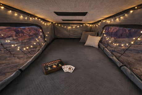 Cozy RV rental interior for glamping. String lights illuminate a comfy bed, scenic views, and Campfire Stories with playing cards.