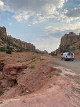 White van, possibly an RV rental, on a winding desert road within a canyon of towering red rock cliffs under cloudy skies.