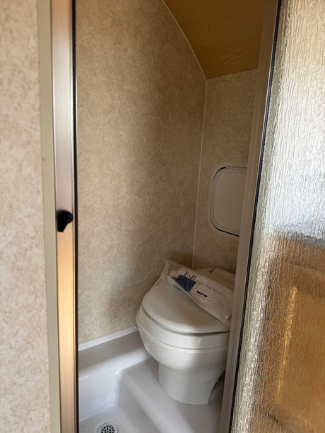 Compact RV rental wet bath with toilet inside shower stall, seen through a partially open frosted glass door.