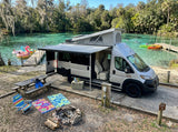 An rv rental (pop-top van) under an awning by a clear spring. A campsite setup includes a picnic table, campfire, and water fun.