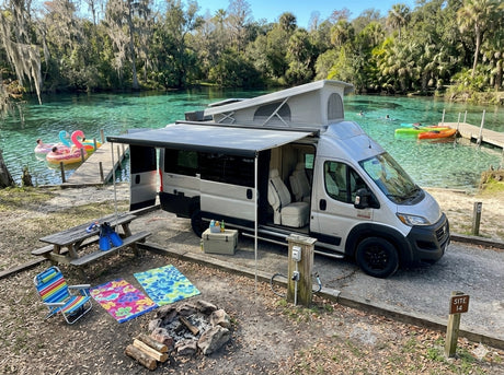 An rv rental (pop-top van) under an awning by a clear spring. A campsite setup includes a picnic table, campfire, and water fun.