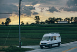 A white commercial van for solar energy drives on a rural road, passing green fields and a power pole. Perfect for an RV rental drive.