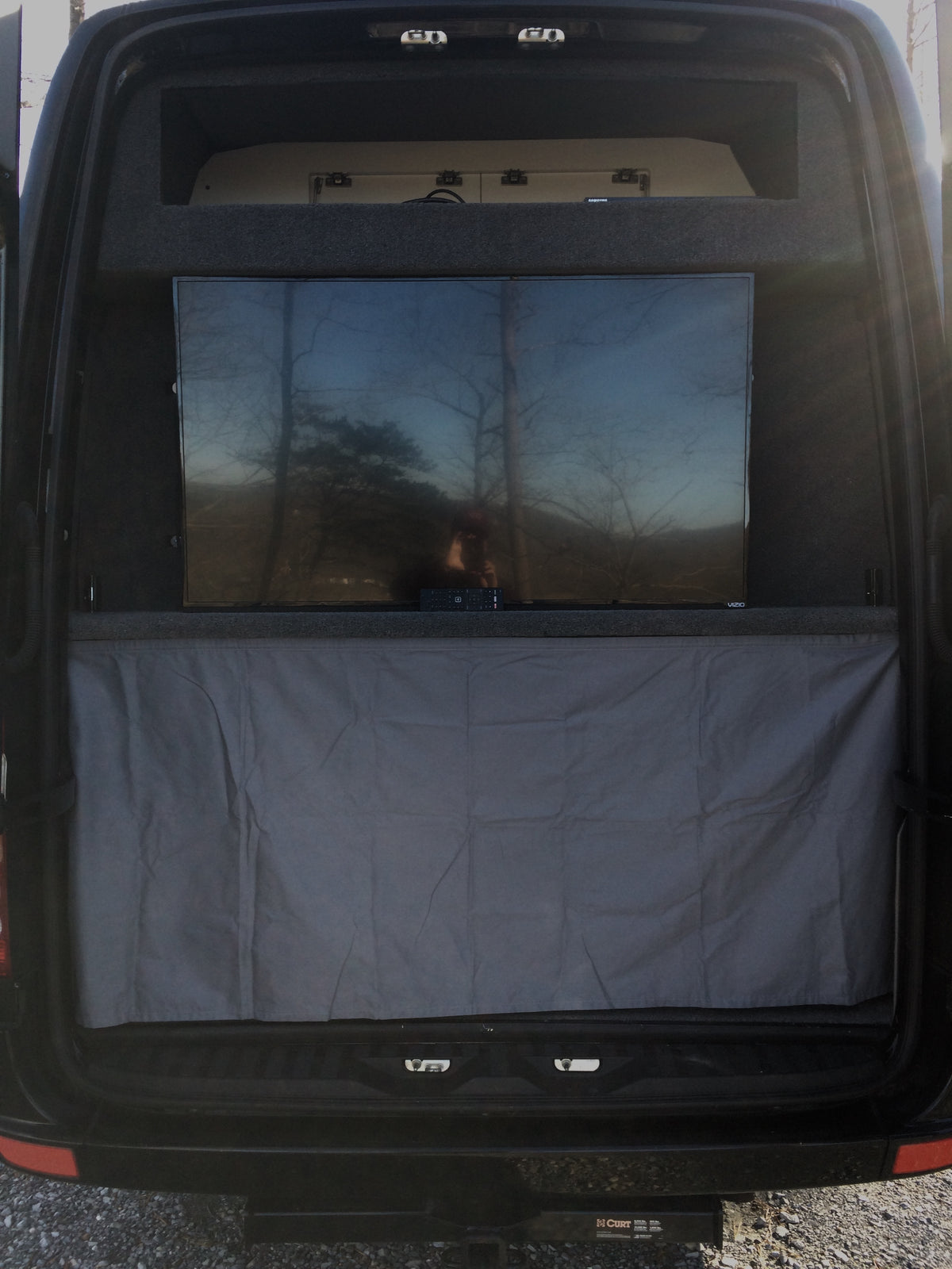 Inside an RV rental: rear view with a large TV reflecting trees, a remote below, and grey curtain. Upper storage and a Curt hitch are visible. — photo 7