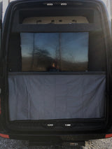 Inside an RV rental: rear view with a large TV reflecting trees, a remote below, and grey curtain. Upper storage and a Curt hitch are visible.