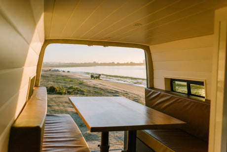 Warm light fills this RV rental's dining area & leather benches, overlooking a serene beach with a parked vehicle.