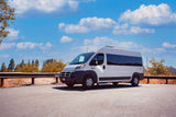 A white Ram ProMaster RV rental van is parked on a scenic road with a bright blue sky and clouds, perfect for travel adventures.