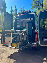 This RV rental shows an adventure-ready van with a mountain bike mounted on a rear slide-out rack, parked in a sunny forest.