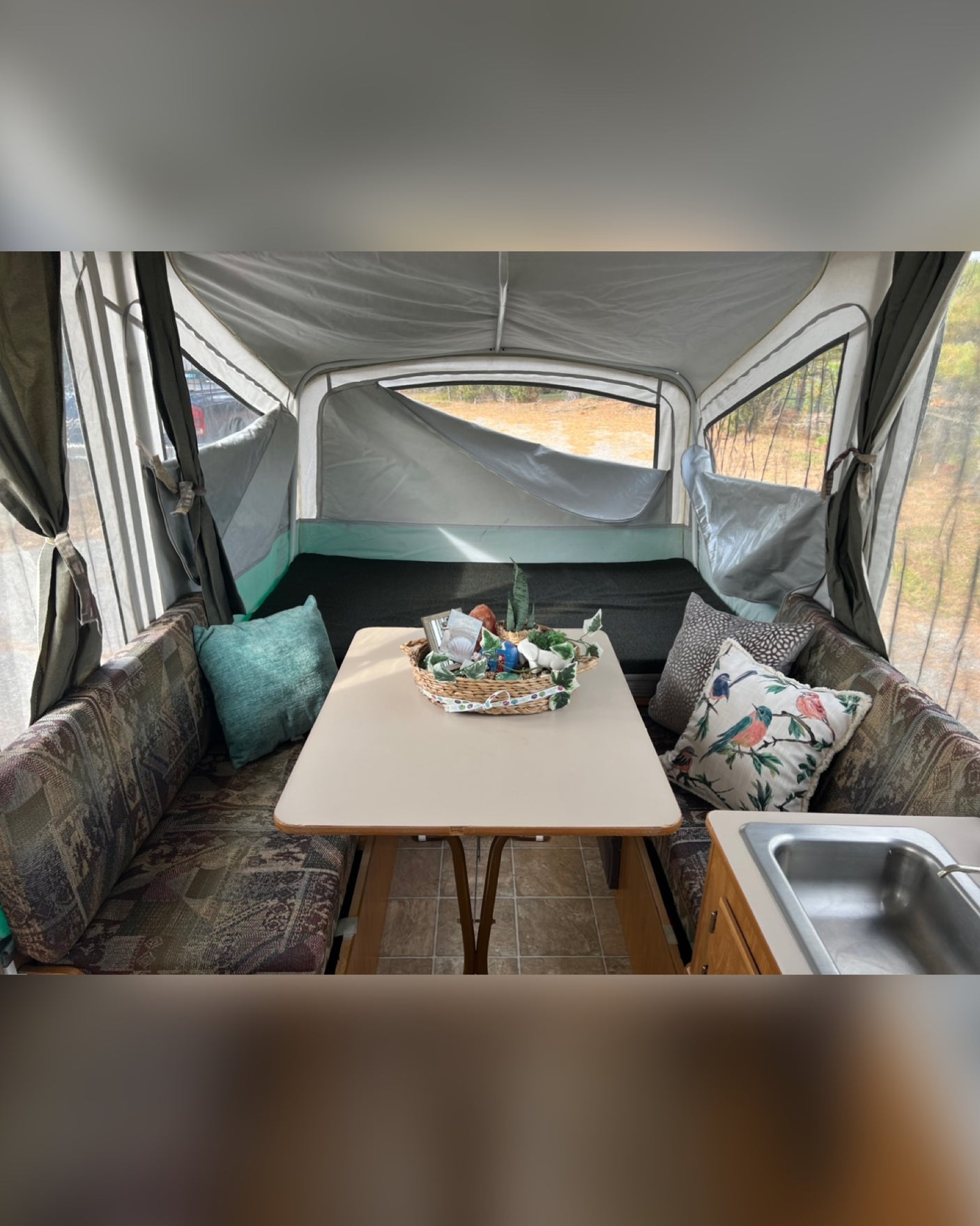 Spacious RV rental interior featuring a bright dining table, patterned benches with pillows, and scenic window views. Includes a sink.