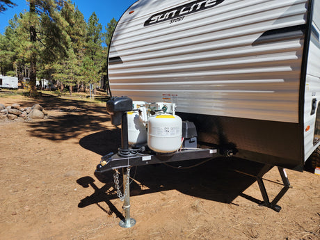 An RV rental, a white Sun Lite Sport travel trailer, parked in a scenic treed campground with visible propane tanks and hitch.