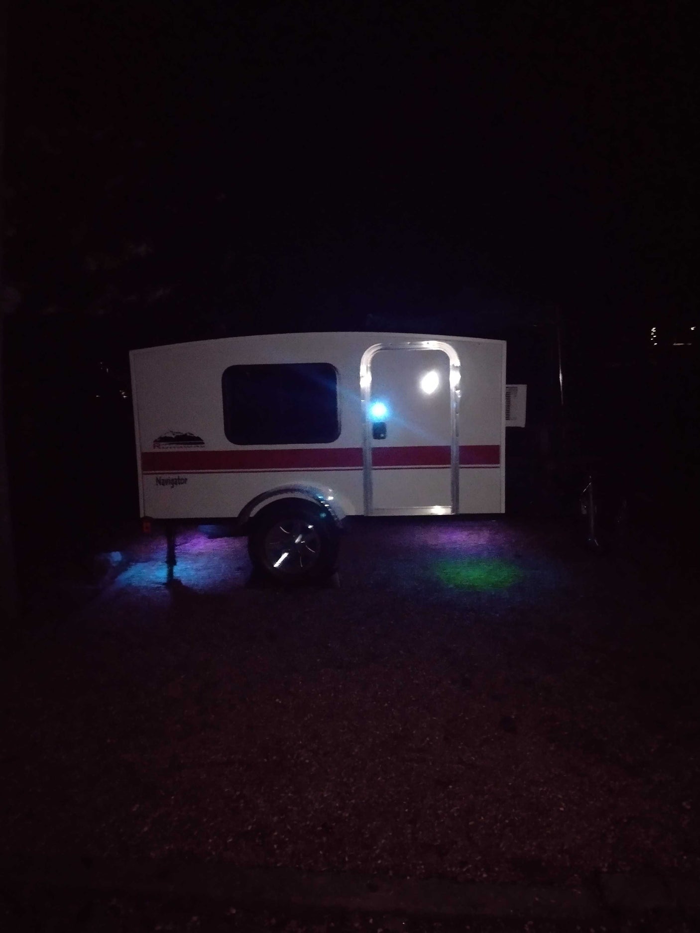 A white RV rental camper glows at night, illuminated by a bright door light and colorful purple, blue, & green ground lights.