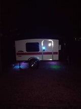 A white RV rental camper glows at night, illuminated by a bright door light and colorful purple, blue, & green ground lights.