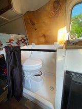 RV rental's compact bathroom: composting toilet in shower pan. Features a wooden cover, curtain, and nearby kitchen.