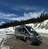 Explore winter roads! This gray Ram ProMaster RV rental is parked on a snowy mountain road, surrounded by evergreen forests.