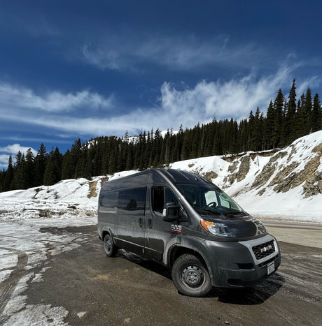 Explore winter roads! This gray Ram ProMaster RV rental is parked on a snowy mountain road, surrounded by evergreen forests.