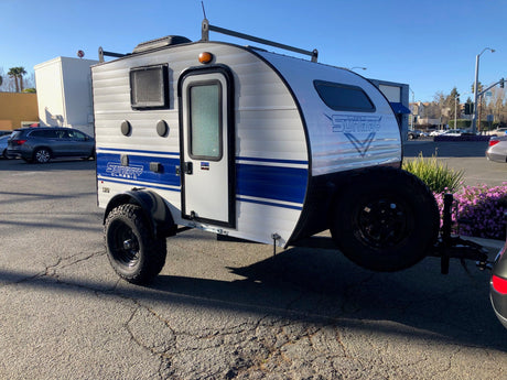 Classic Sunray teardrop RV rental: a compact, light grey travel trailer with blue accents and rugged tires, parked outdoors.