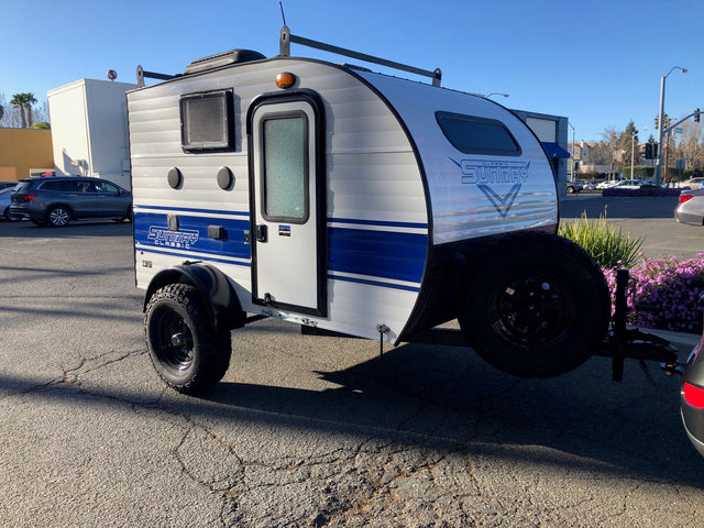 Classic Sunray teardrop RV rental: a compact, light grey travel trailer with blue accents and rugged tires, parked outdoors.