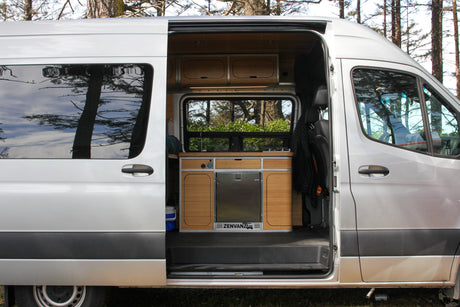 Silver RV rental: open door reveals custom Zenvanz camper interior with kitchenette, fridge, and cabinets. Ready for adventure!