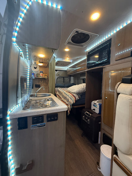 Inside an RV rental, featuring a compact kitchen with stove and sink, cozy sleeping area, microwave, and bright LED lighting.