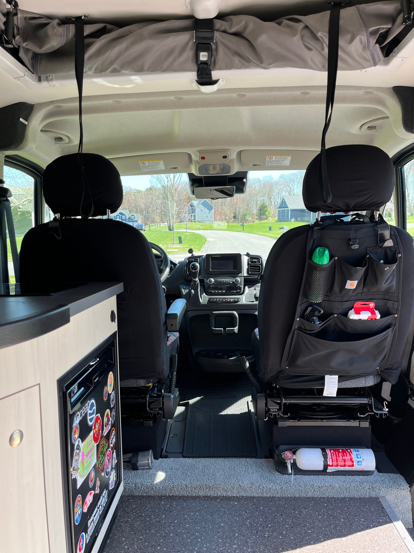 Inside an RV rental, looking forward. Features driver's cabin, front seats with storage, dashboard, fridge, and safety fire extinguisher.