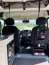 Inside an RV rental, looking forward. Features driver's cabin, front seats with storage, dashboard, fridge, and safety fire extinguisher.