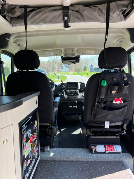 Inside an RV rental, looking forward. Features driver's cabin, front seats with storage, dashboard, fridge, and safety fire extinguisher.