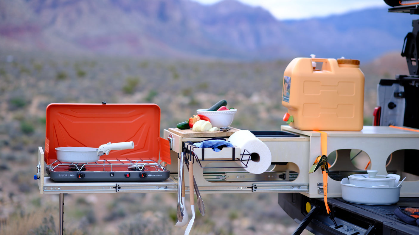 RV rental outdoor kitchen with a camping stove, fresh vegetables, water container, and cooking gear in a desert setting.