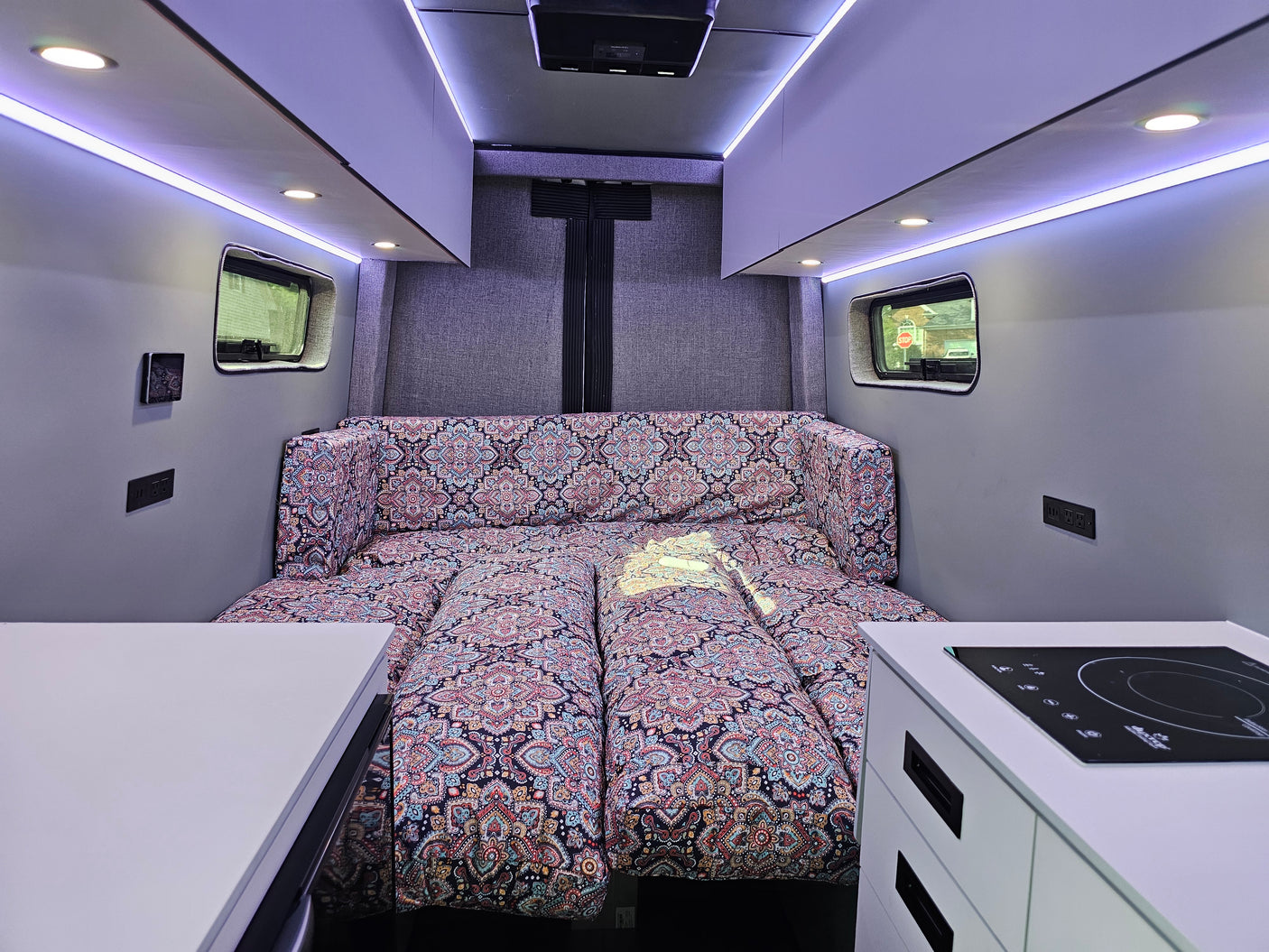Cozy RV rental interior features a vibrant patterned bed, sleek induction cooktop, and bright LED strip lighting. Ideal for road trips.