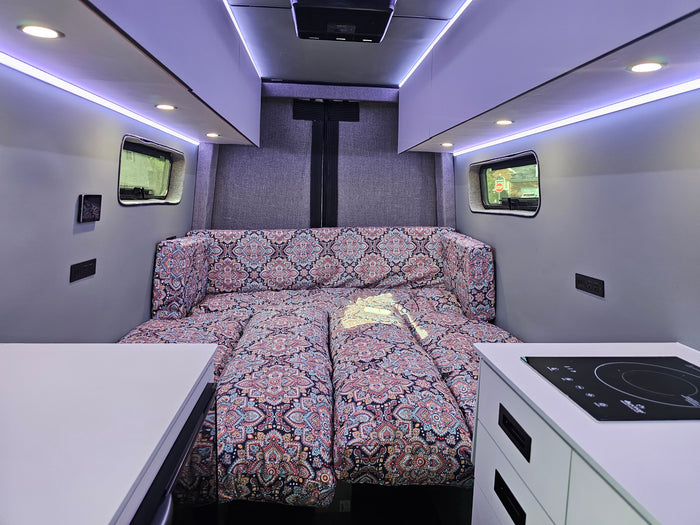 Cozy RV rental interior features a vibrant patterned bed, sleek induction cooktop, and bright LED strip lighting. Ideal for road trips.