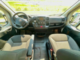 Inside an RV rental: driver's perspective of the cab, dashboard, steering wheel (Ram logo), and seats, looking out to a sunny field.