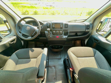 Inside an RV rental: driver's perspective of the cab, dashboard, steering wheel (Ram logo), and seats, looking out to a sunny field.